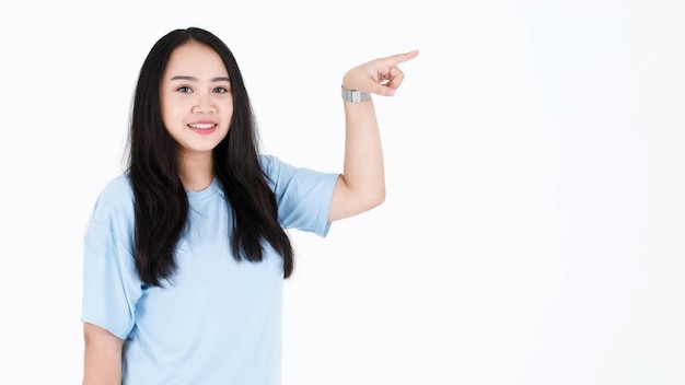 Portrait studio shot of Asian young chubby plump long black hair female model in blue casual t shirt and wristwatch holding hand pointing blank space showing presenting product on white background.