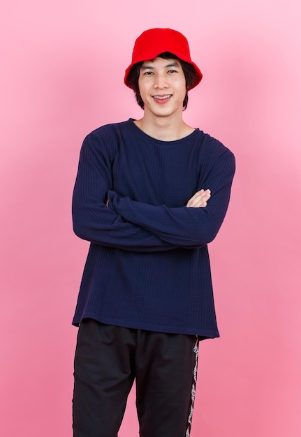 Portrait studio shot Asian young urban teen fashion male model in street style outfit long sleeve shirt red bucket hat standing crossed arms smiling look at camera on pink background