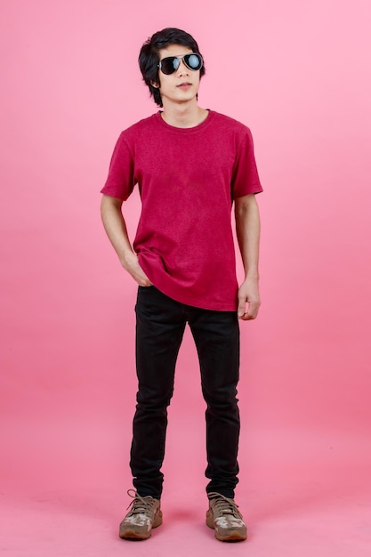 Portrait studio shot of Asian young urban teenager fashion male model in casual street style outfit t shirt and black jeans with aviator sunglasses standing posing look at camera on pink background