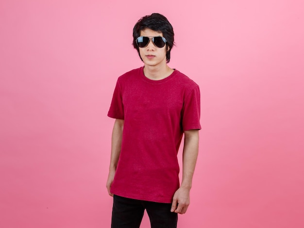 Portrait studio shot of Asian young urban teenager fashion male model in casual street style outfit t shirt and black jeans with aviator sunglasses standing posing look at camera on pink background