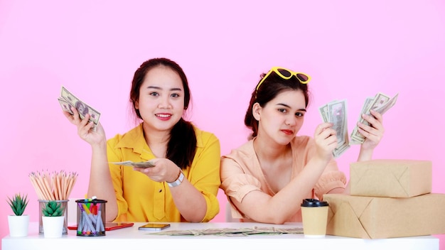 Portrait studio shot of two Asian rich wealthy female startup small business entrepreneur businesswoman partner sitting together holding full hundred dollar banknotes in hands on pink background.