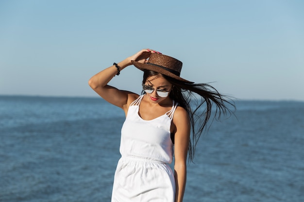 Portrait of stunning dark-haired girl in white dress, sunglasses and brown hat near the sea on a sunny day. .