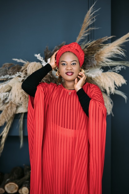 Portrait of stunning pretty African woman with in red dress and traditional shawl on her head, posing on dark studio background with stylish boho decorations.