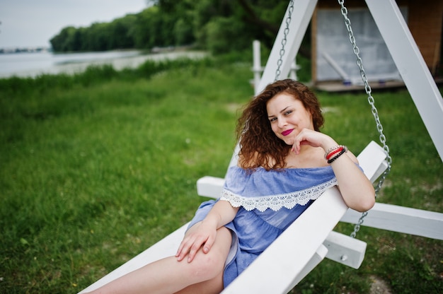 Portrait of a stunning young woman wearing blue marine-styled dress sitting on swings on the lakeside.