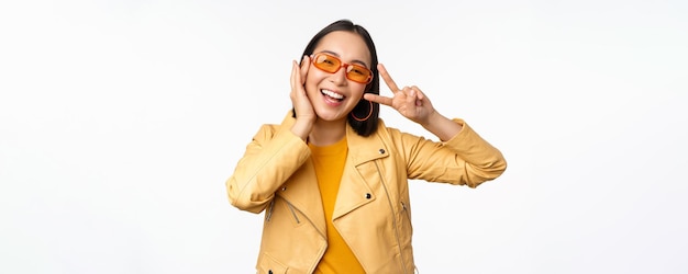 Portrait of stylish asian modern girl wearing sunglasses and yellow jacket showing peace vsign gesture standing over white background happy smiling face