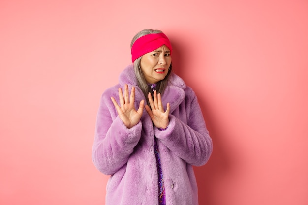 Portrait of stylish asian senior woman in purple winter coat, cringe from something disgusting, frowning and turn away awkward, showing stop gesture, refusing on pink background