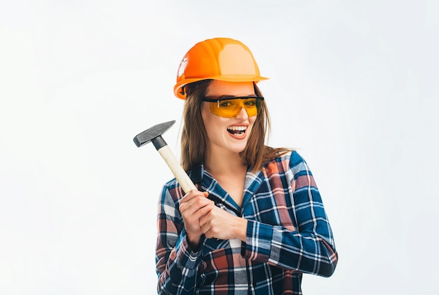 Portrait of stylish beautiful young woman with standing on white background Woman wearing helmet and glasses Smiling sexy young engineer holding a hammer