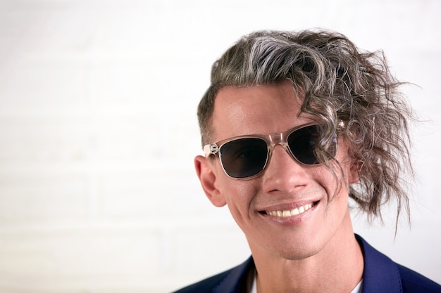 Portrait of stylish businessman with curly long hair in sunglasses smiling at the camera on white background