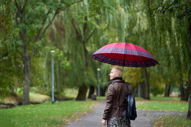 Portrait of stylish cute short haired blonde woman walking in the autumn park with red umbrella View from back