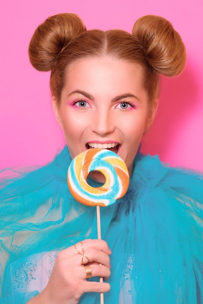 Portrait of stylish cute young blonde woman with colorful lollipop on pink background