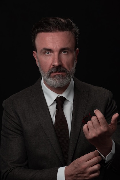 Portrait of a stylish elegant senior businessman with a beard and casual business clothes in photo studio isolated on dark background adjusting the suit