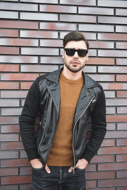 Portrait of stylish handsome young man with bristle standing outdoors and leaning on brick wall