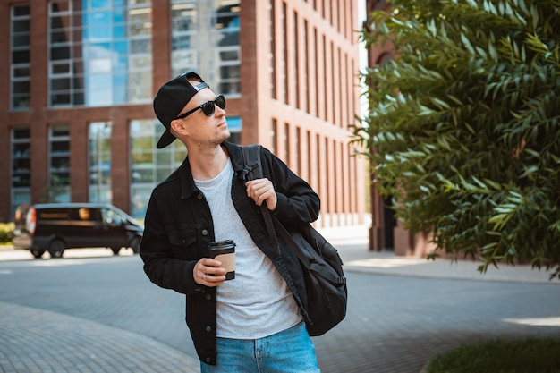 Portrait of a stylish hipster guy in sunglasses with a backpack in the city against the background of a modern city building