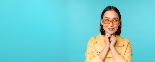 Portrait of stylish korean woman in sunglasses smiling and looking aside at logo coquettish standing over blue background Copy space