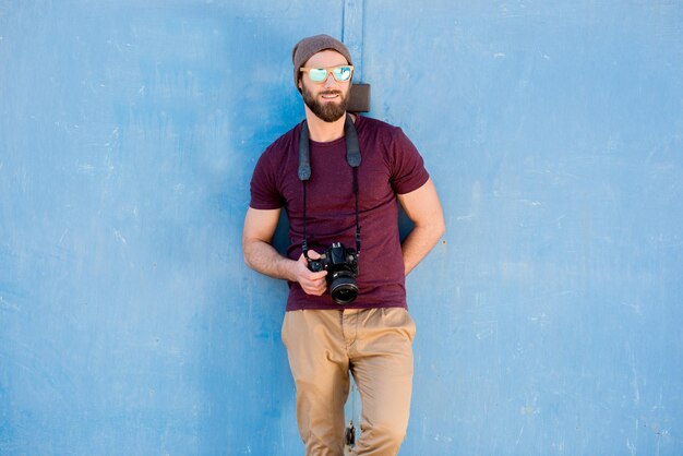 Photo portrait of a stylish photographer dressed casual in t-shirt and hat standing with camera on the blue background