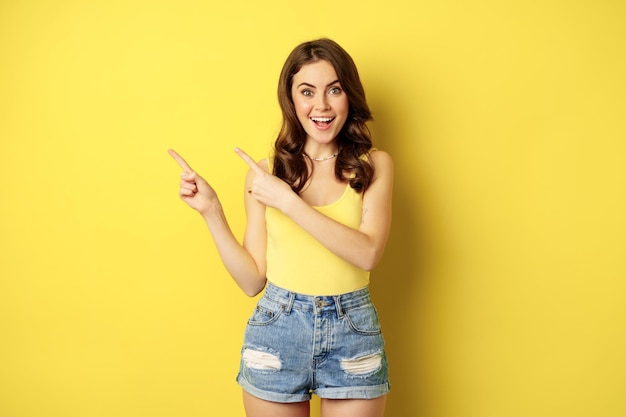 Portrait of stylish summer girl, brunette woman showing way, pointing fingers left, advertising, standing over yellow background. Copy space