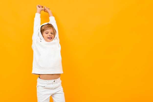 Portrait of stylish young caucasian boy with medium fair hair in white hoody, black trousers isolated on yellow background