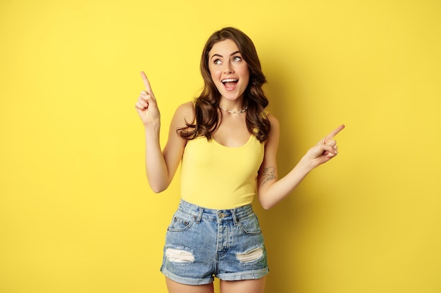 Portrait of stylish young female model, showing advertisement, pointing at logo or banner, demonstrating two ways, choices in store, standing over yellow background