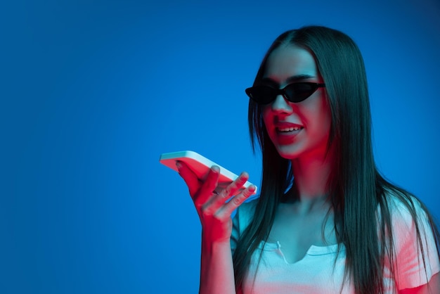 Portrait of stylish young girl recording voice message on phone posing isolated over blue studio background in neon light