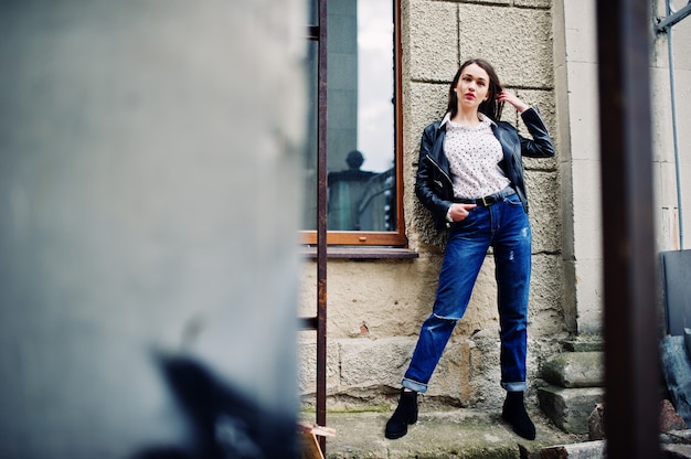 Photo portrait of stylish young woman wearing on leather jacket and ripped jeans at streets of city.