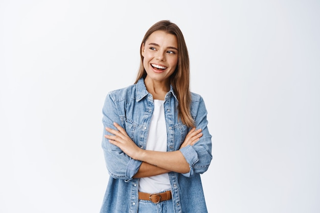 Portrait of stylish young woman with blond hair, cross arms on chest, looking left at empty space for banner and smiling pleased, checking out advertisement, white wall