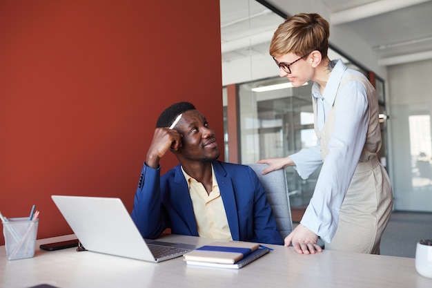 Portrait of successful African-American businessman looking at female colleague and smiling while working in modern office interior with red wall, copy space