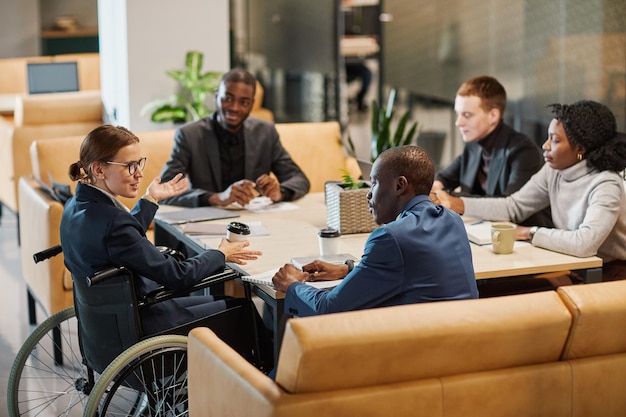 Portrait of successful businesswoman using wheelchair at meeting table and talking to diverse group of colleagues in modern office space