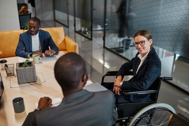 Portrait of successful businesswoman using wheelchair at meeting and talking to group of colleagues in modern office space