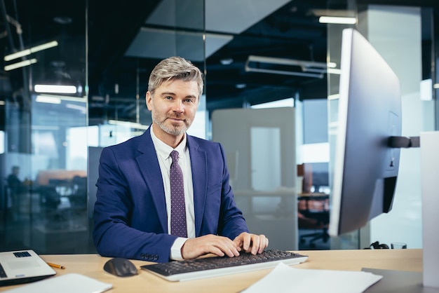 Portrait of a successful grayhaired man with a beard businessman working at a computer in a modern office and looking at the camera