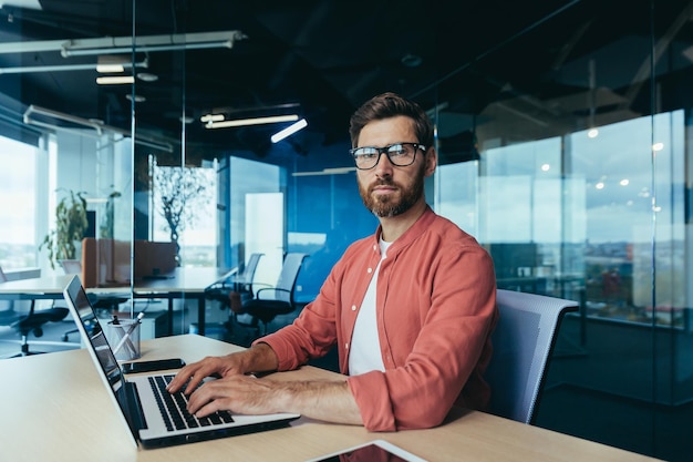 Portrait of a successful programmer inside the office a man with a beard glasses and a red shirt