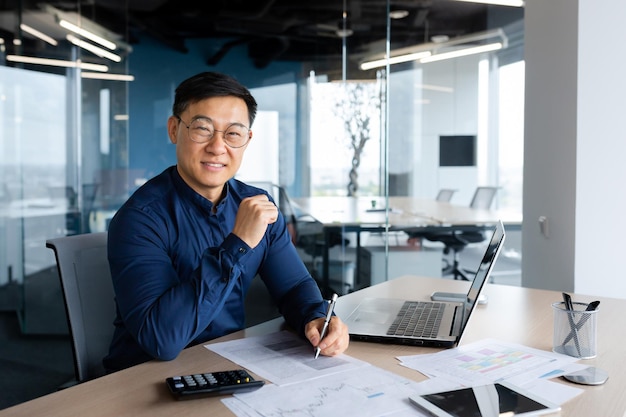 Portrait successful satisfied asian financier accountant man in shirt and glasses working inside