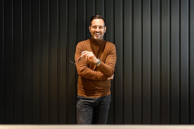 Photo portrait of successful smiling businessman holding eyeglasses and posing confidently wall office