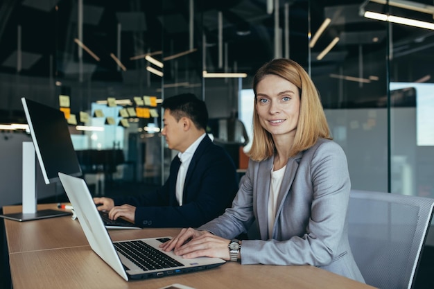 Portrait of successful woman business team member business woman looking at camera and smiling working on laptop in modern office