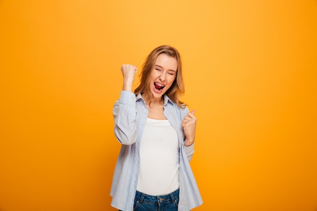 Portrait of successful woman wearing braces yelling and clenching fists, acting like winner or lucky person, isolated over yellow background