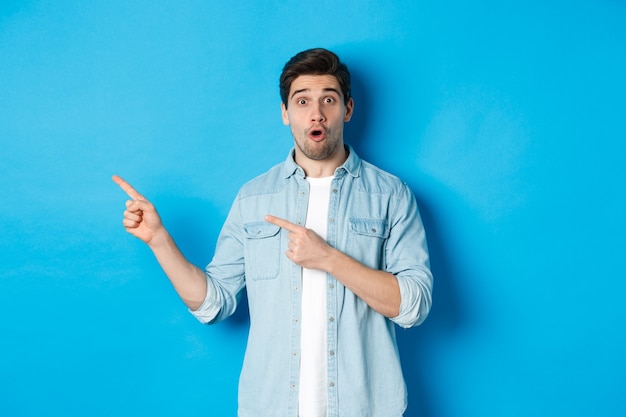 Portrait of surprised adult man in casual clothes showing announcement, pointing fingers left and looking amazed, standing against blue wall