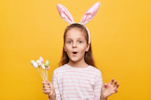 Portrait of surprised cute little girl with rabbit ears with cake pops in hands looking at camera with amazement saying wow posing isolated on yellow background