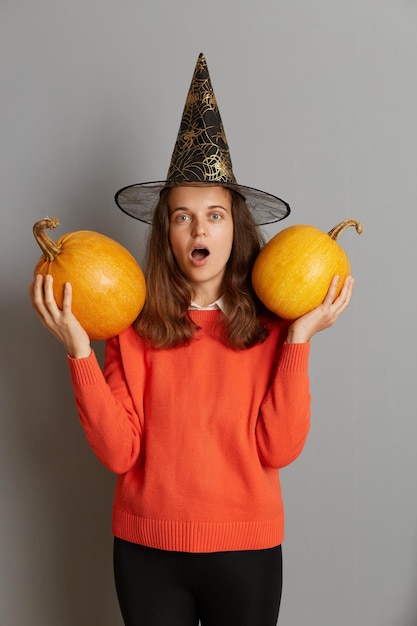 Portrait of surprised little scared Caucasian girl standing with pumpkins in her handswearing witch hat posing with Halloween pumpkin standing on gray background looking at camera with open mouth