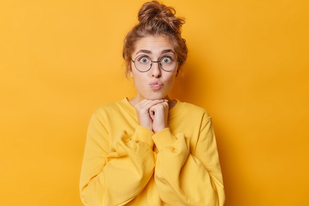 Portrait of surprised lovely woman keeps lips rounded hands under chin has dark hair gathered in bun wears round spectacles and jumper isolated over yellow background Human face expressions