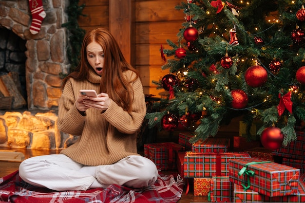 Portrait of surprised redhead young woman wearing winter sweater getting good news on mobile phone