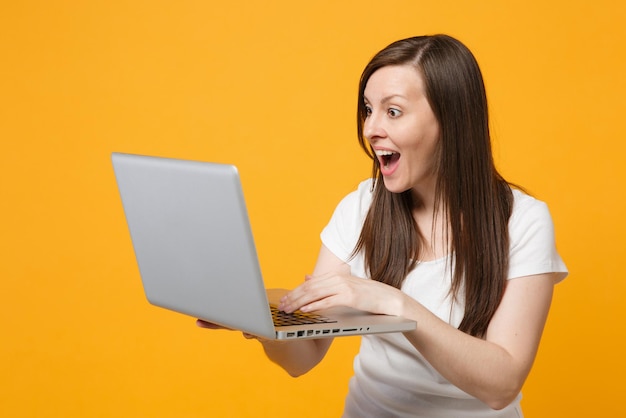 Portrait of surprised young woman in white casual clothes keeping mouth open, using laptop pc computer isolated on bright yellow orange wall background. People lifestyle concept. Mock up copy space.