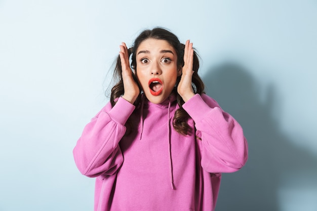 Portrait of surprised young woman with two ponytails wondering and grabbing her head over blue in studio