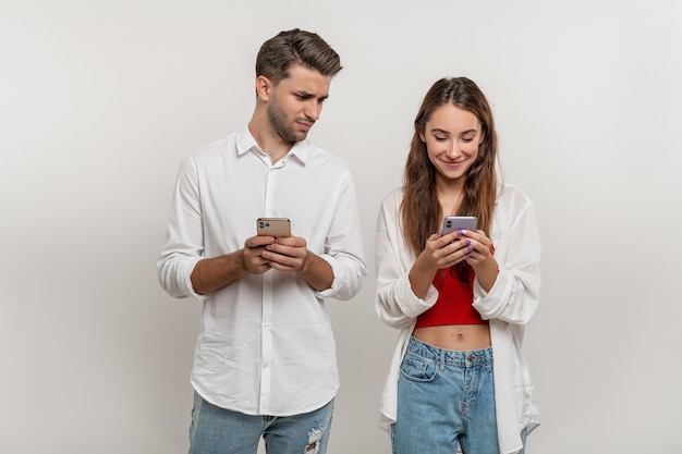 Portrait of suspecting man spying on his happy girlfriend using mobile phone isolated over white