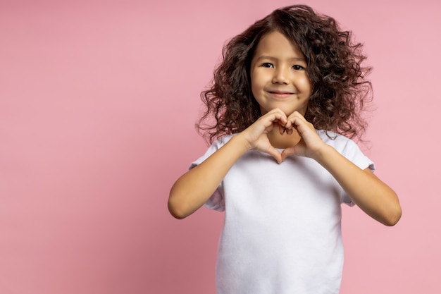 Portrait of sweet cheerful girl wearing white t shirt, having dark curly hair, showing heart gesture, looking aside smiling, standing isolated.