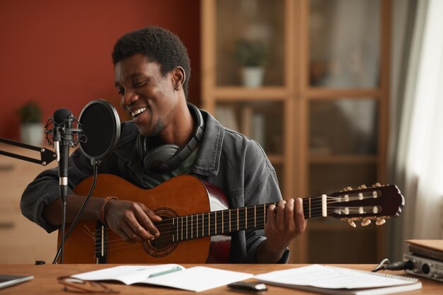 Photo portrait of talented african-american man singing to microphone and playing guitar while recording music in studio, copy space