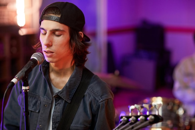 Portrait of talented young musician singing to microphone with eyes closed in colored studio lights