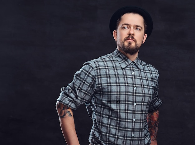 Portrait of a tattoed middle age hipster man with beard and hairstyle dressed in a checkered shirt and hat, pose in a studio. Isolated on a dark textured background.