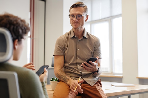 Photo portrait of tattooed adult man talking to colleague while sitting on desk in office copy space