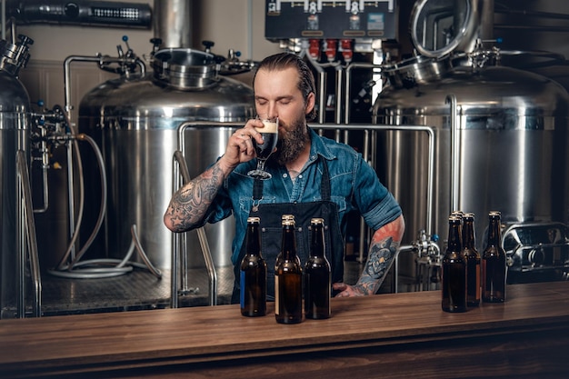 Portrait of tattooed, bearded hipster male manufacturer tasting beer in the microbrewery.