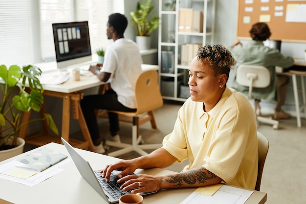 Portrait of tattooed black woman using laptop in office while working with team in background copy s