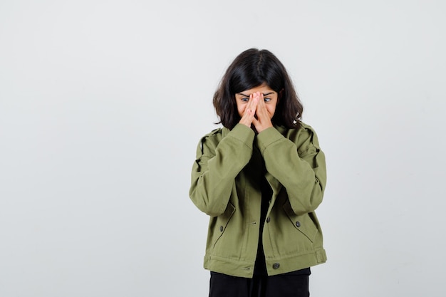 Portrait of teen girl holding hands on face in army green jacket and looking anxious front view